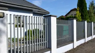 Nowoczesne ogrodzenie murowane tynkowane z metalowymi przęsłami, otaczające zadbany dom z ogrodem.