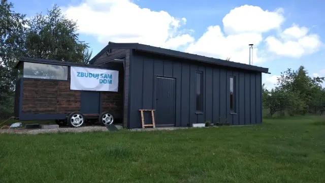 Mobilny garaż bez pozwolenia, z banerem "Zbuduj sam dom", stoi na trawie.